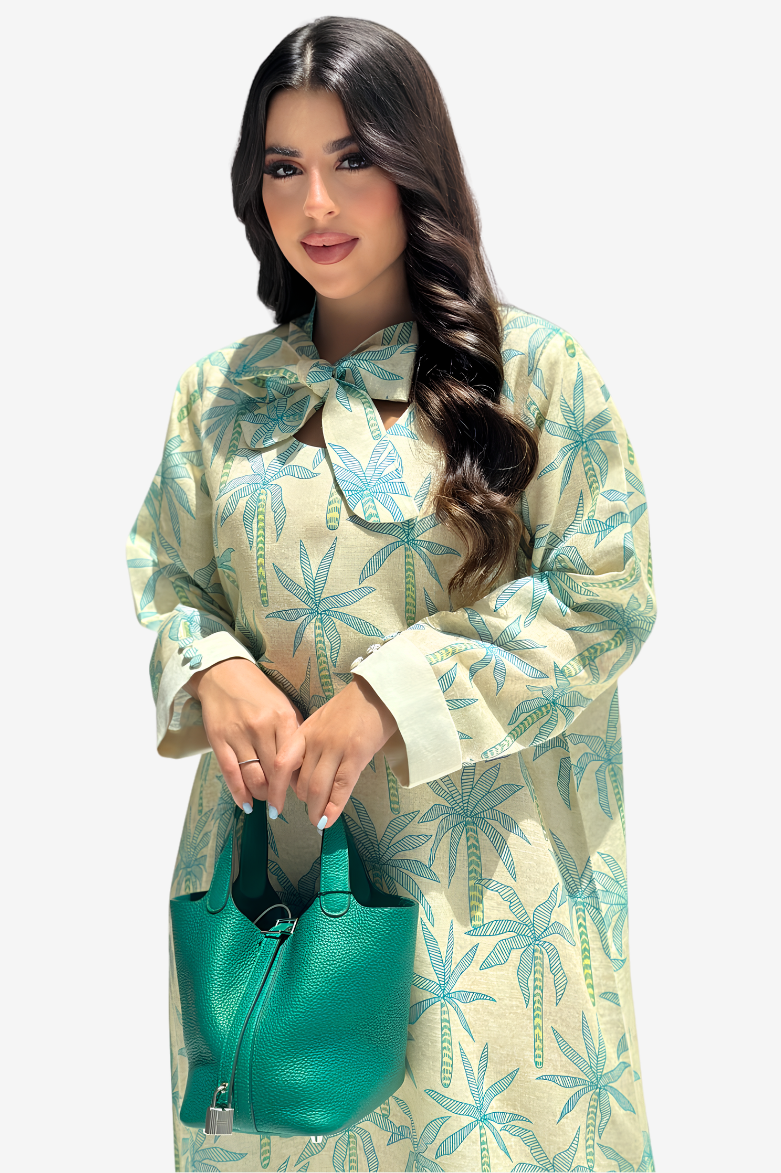 A woman modeling a long-sleeve cream-colored dress with a teal palm tree pattern, holding a matching teal handbag against a light gray background.