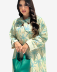 A woman modeling a long-sleeve cream-colored dress with a teal palm tree pattern, holding a matching teal handbag against a light gray background.