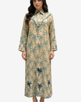 A woman modeling a long-sleeved beige maxi dress with a blue palm tree pattern, standing against a light gray background.