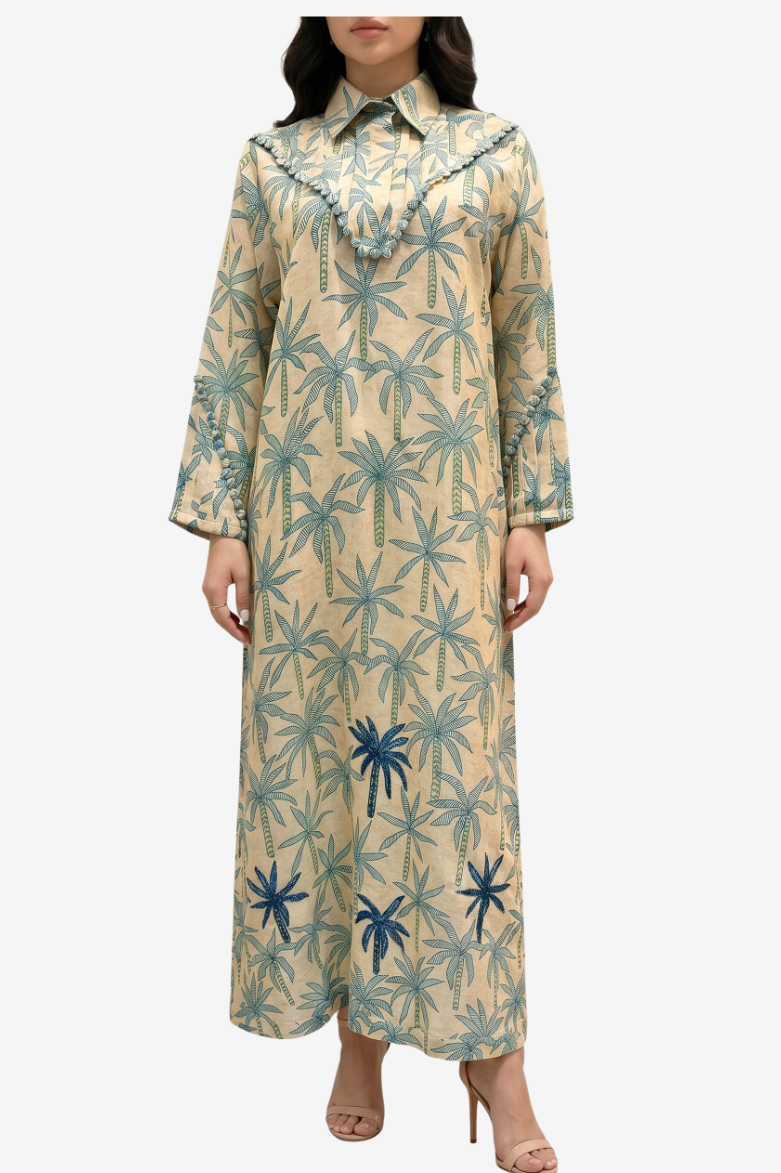 A woman modeling a long-sleeved beige maxi dress with a blue and green palm tree pattern, featuring a ruffled collar and buttoned cuffs, set against a plain light gray background.