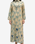 A woman modeling a long-sleeved beige maxi dress with a blue and green palm tree pattern, featuring a ruffled collar and buttoned cuffs, set against a plain light gray background.