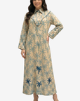 A woman modeling a long-sleeved beige maxi dress with a blue palm tree pattern, standing against a light gray background.