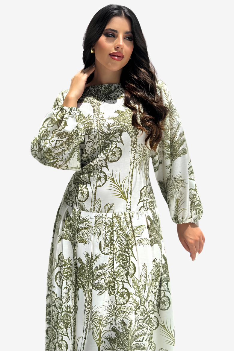 A woman modeling a long-sleeved maxi dress with a green botanical and tropical leaf pattern on a white background, set against a plain light gray studio backdrop.