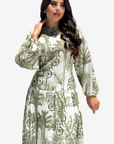 A woman modeling a long-sleeved maxi dress with a green botanical and tropical leaf pattern on a white background, set against a plain light gray studio backdrop.