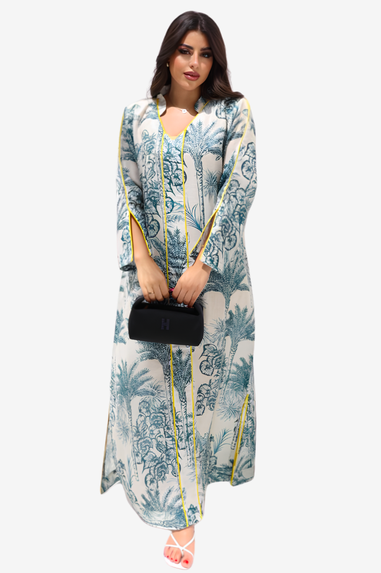 A woman modeling a long-sleeved maxi dress with a blue and green botanical print on a white base, featuring yellow piping details, holding a small black handbag against a light gray background.