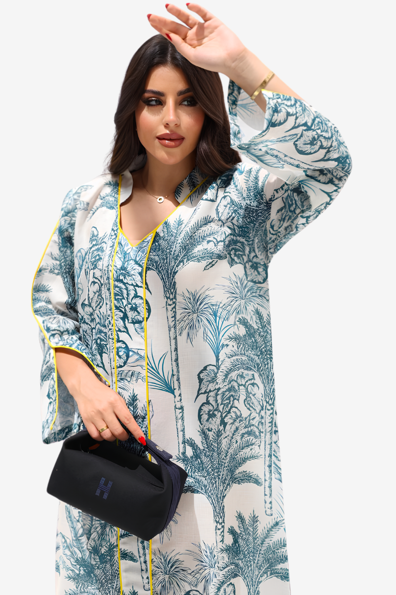 A woman modeling a long-sleeve white kaftan-style tunic with a blue tropical palm tree print and yellow piping trim, holding a black handbag against a light gray studio background.