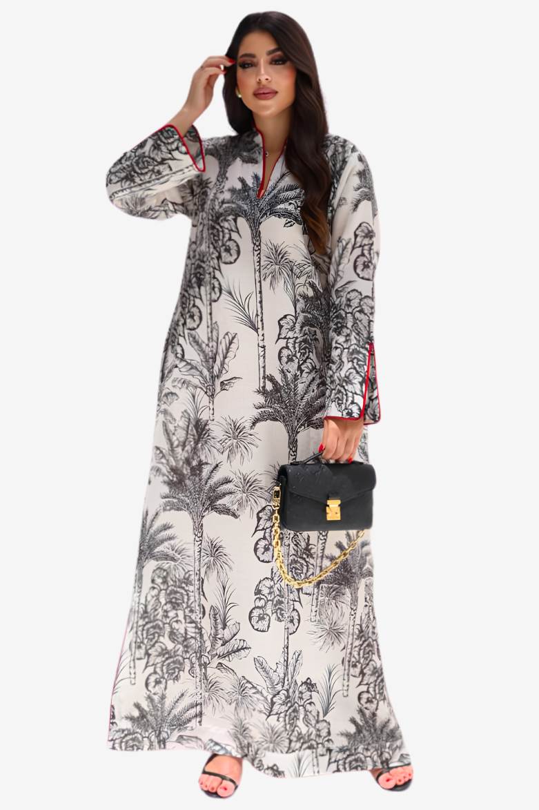 A woman modeling a long-sleeved, floor-length kaftan dress with a black and white botanical print, holding a black handbag with a gold chain strap against a light gray background.