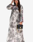 A woman modeling a long-sleeved, floor-length kaftan dress with a black and white botanical print, holding a black handbag with a gold chain strap against a light gray background.