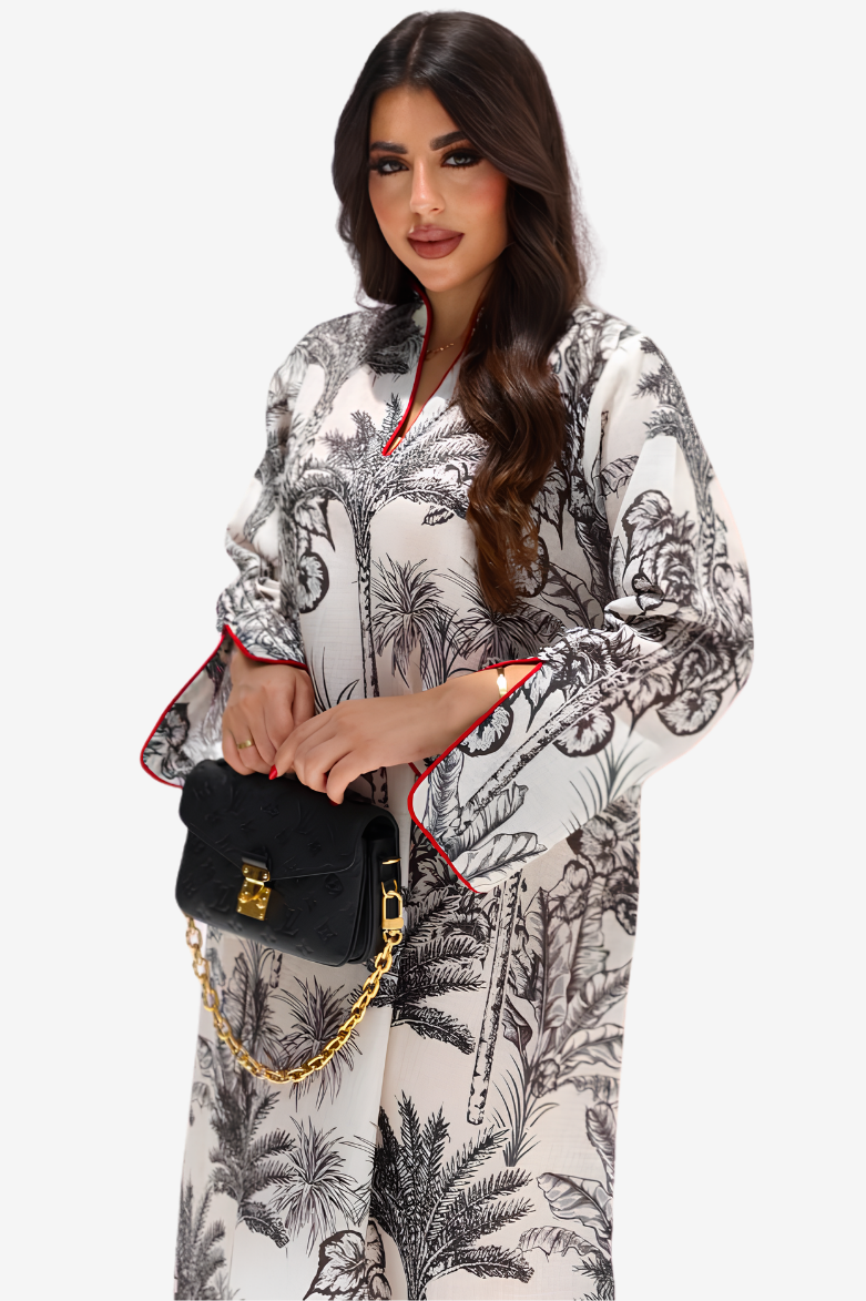 A woman modeling a long-sleeved white kaftan-style dress with a black botanical and palm tree print, featuring red piping along the collar and cuffs. She is holding a black handbag with a gold chain strap against a plain light gray background.