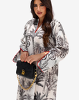 A woman modeling a long-sleeved white kaftan-style dress with a black botanical and palm tree print, featuring red piping along the collar and cuffs. She is holding a black handbag with a gold chain strap against a plain light gray background.