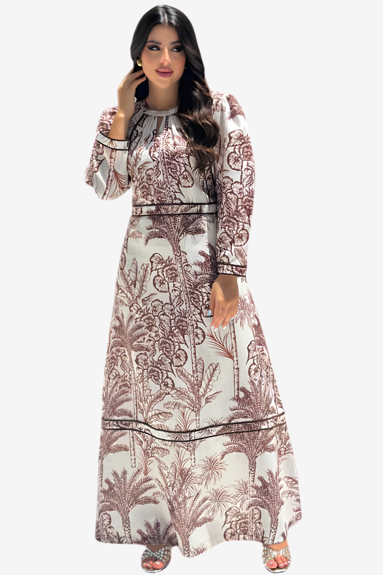 A woman modeling a long-sleeved maxi dress with a brown palm tree and floral pattern on a cream background, set against a plain light gray studio background.