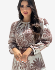 A woman modeling a long-sleeved dress with a brown botanical and palm tree print on a light beige background, set against a plain light gray studio background.