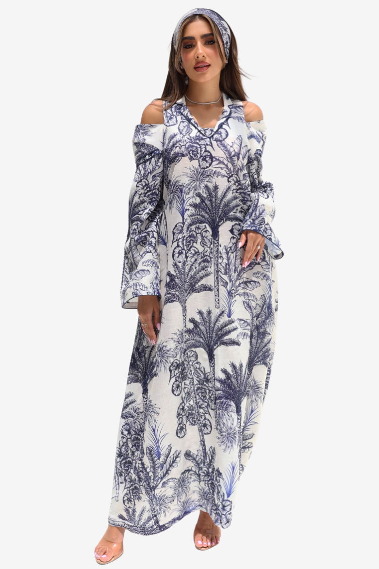 A woman modeling a long cold-shoulder maxi dress with a blue palm tree and floral toile-style print on a white background, paired with a matching headband, against a light gray studio background.