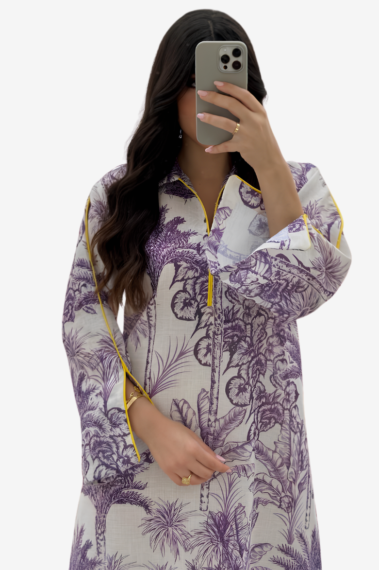 A woman takes a mirror selfie wearing a long-sleeve tunic top with a purple botanical print on a white base, featuring yellow piping details along the collar, cuffs, and front zipper.