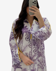 A woman takes a mirror selfie wearing a long-sleeve tunic top with a purple botanical print on a white base, featuring yellow piping details along the collar, cuffs, and front zipper.