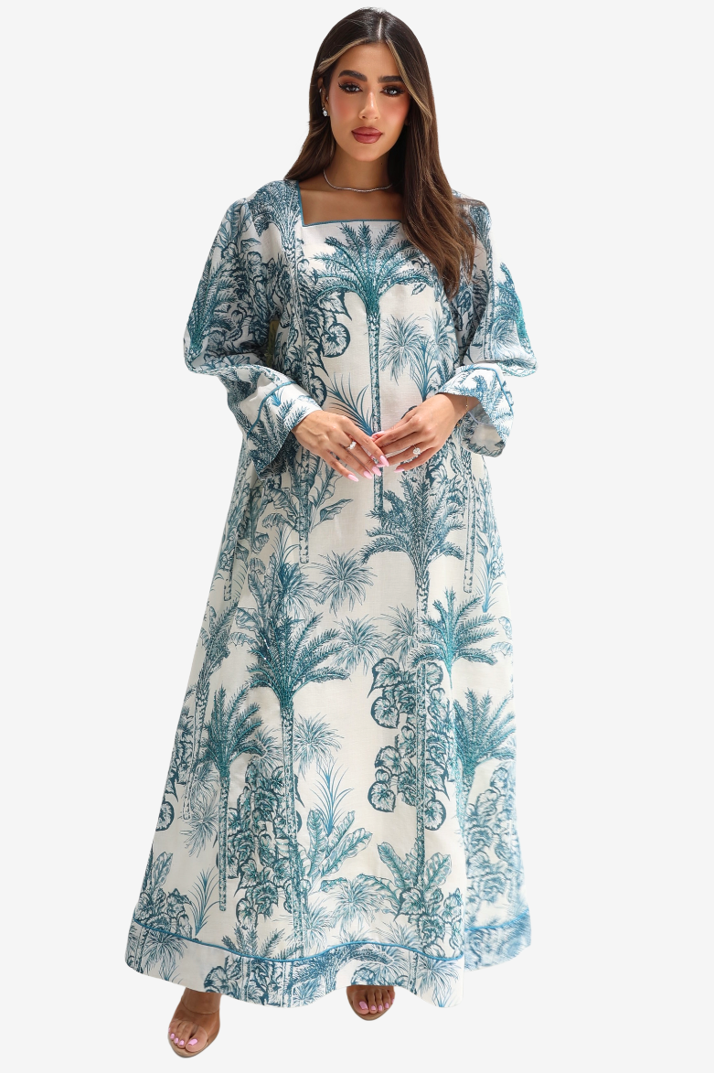 A woman modeling a long-sleeved maxi dress with a blue and white tropical palm tree print, set against a plain light gray background.