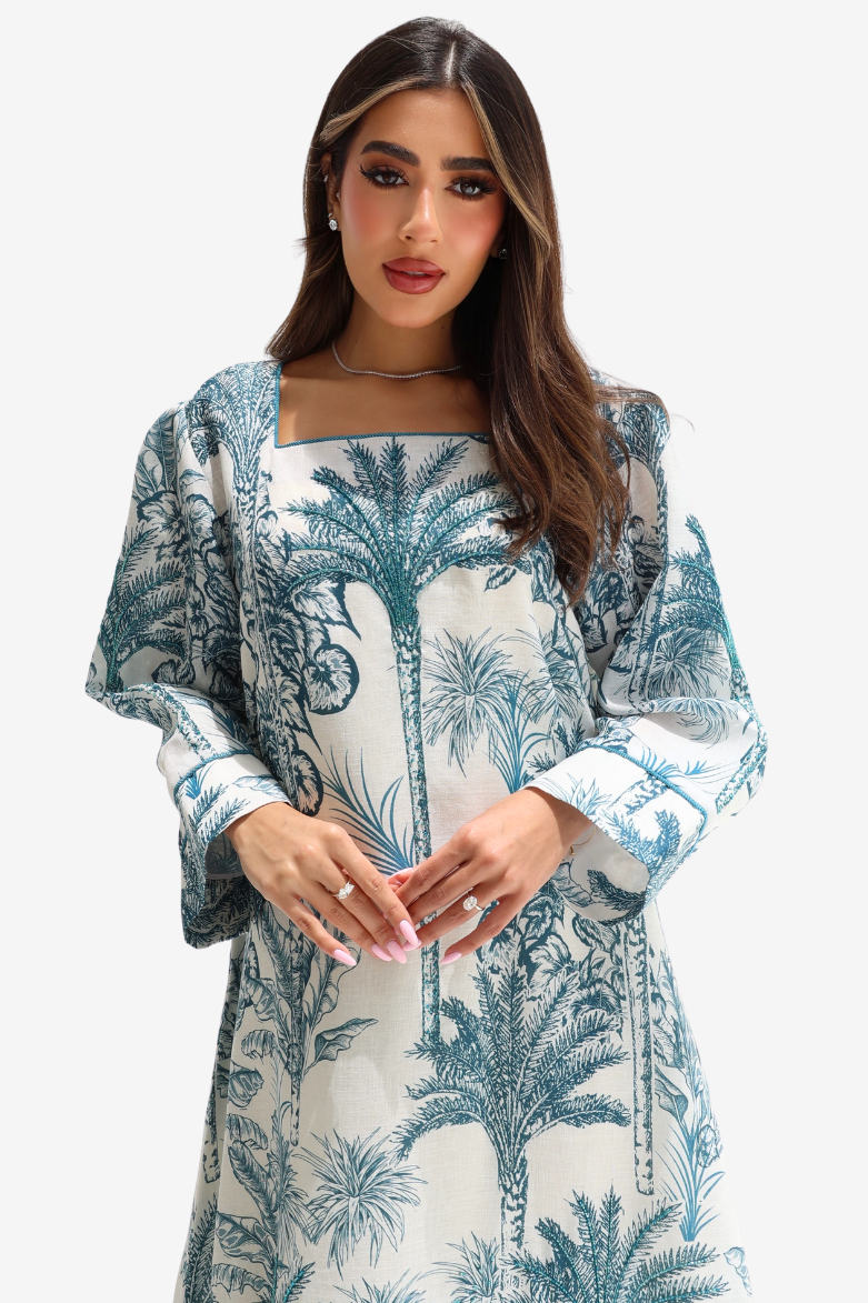 A woman wearing a long-sleeve dress with a blue palm tree toile pattern on a white background, set against a plain light gray studio backdrop.