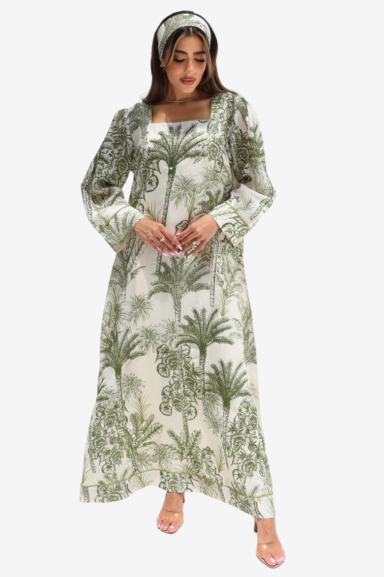 A woman modeling a long-sleeved maxi dress with a green palm tree botanical print on a cream background, paired with a matching headband, against a plain light gray background.