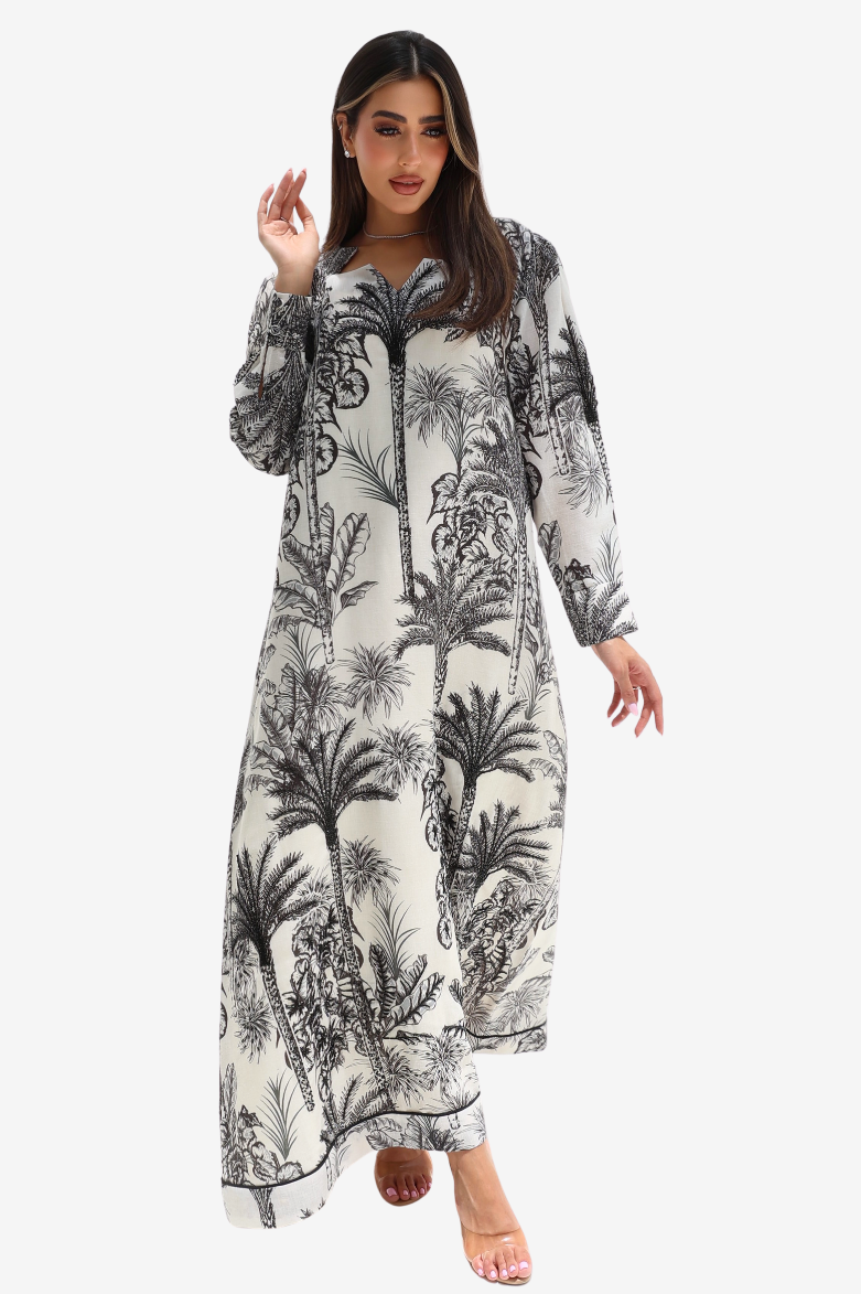 A woman modeling a long-sleeved maxi dress with a black palm tree and botanical print on a warm off-white background, set against a solid light gray studio background.