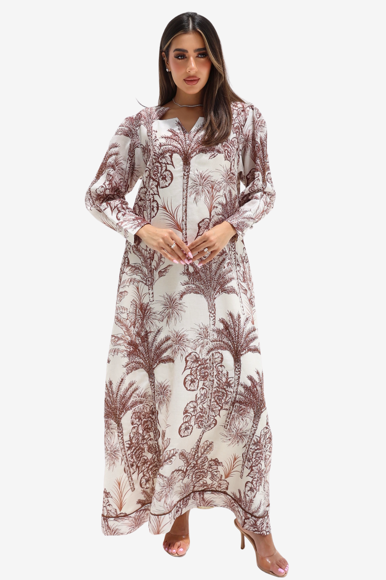 A woman modeling a long-sleeved maxi dress with a brown palm tree and floral toile pattern on a cream background, set against a plain light gray studio background.