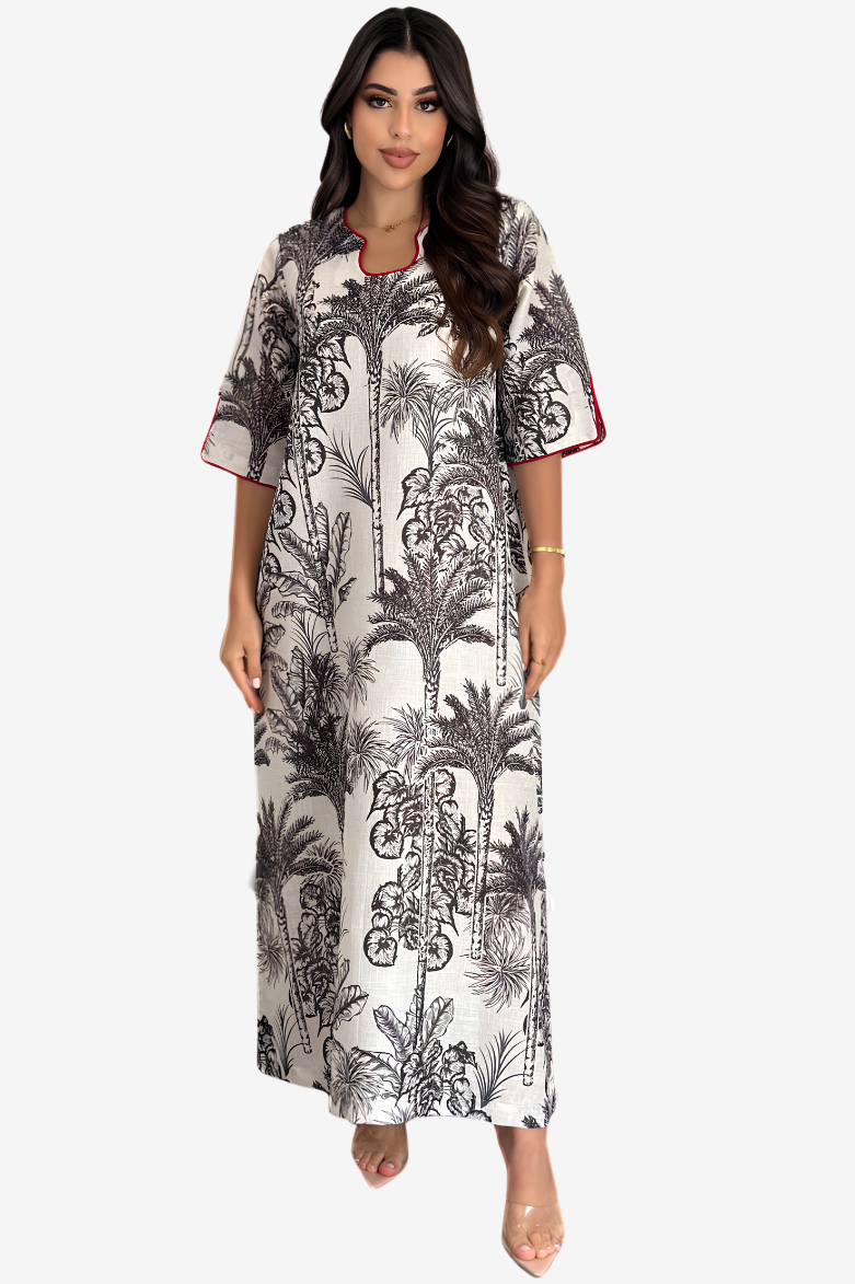 A woman modeling a short-sleeve maxi dress with a black palm tree and tropical leaf print on a white background, featuring red piping details along the collar and sleeve cuffs.