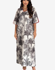 A woman modeling a short-sleeve maxi dress with a black palm tree and tropical leaf print on a white background, featuring red piping details along the collar and sleeve cuffs.