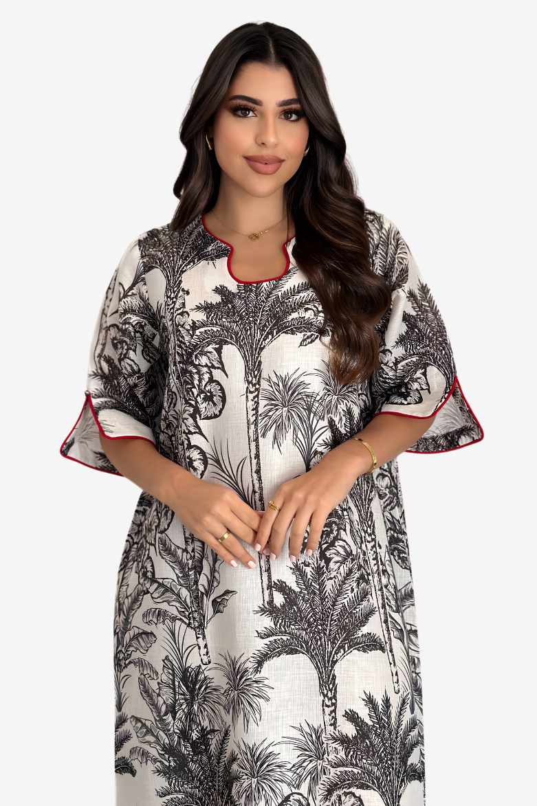 A woman modeling a white kaftan dress with a black palm tree print and red piping trim on the collar and sleeve cuffs, set against a plain light gray background.