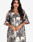 A woman modeling a white kaftan dress with a black palm tree print and red piping trim on the collar and sleeve cuffs, set against a plain light gray background.