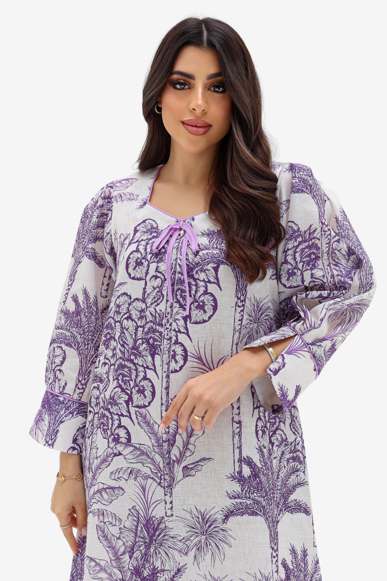 A woman modeling a loose, long-sleeve tunic dress with a purple botanical and palm tree print on a white background, featuring a purple tie-neck detail.
