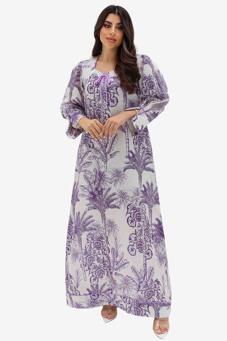 A woman modeling a long-sleeved maxi dress with a purple palm tree toile pattern on a white background, set against a plain light gray studio backdrop.