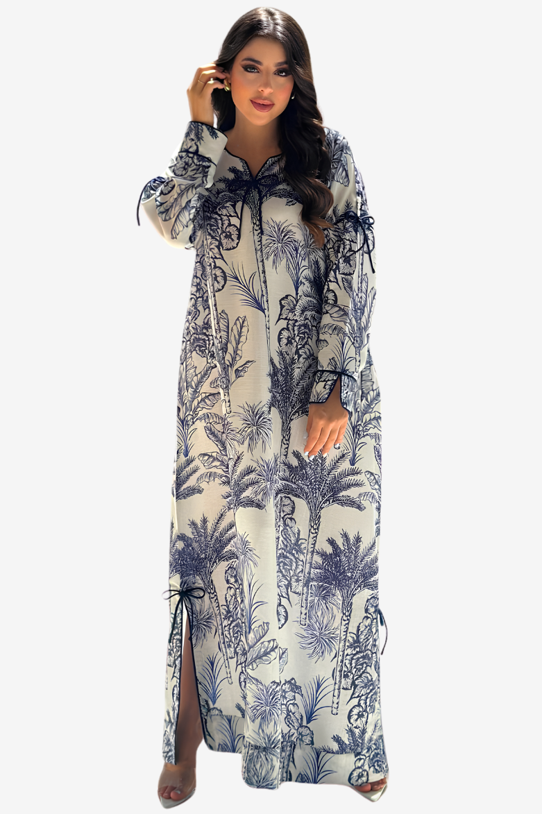 A woman modeling a long-sleeved maxi dress with a blue and navy palm tree pattern on a cream background, set against a plain light gray studio background.