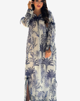 A woman modeling a long-sleeved maxi dress with a blue and navy palm tree pattern on a cream background, set against a plain light gray studio background.