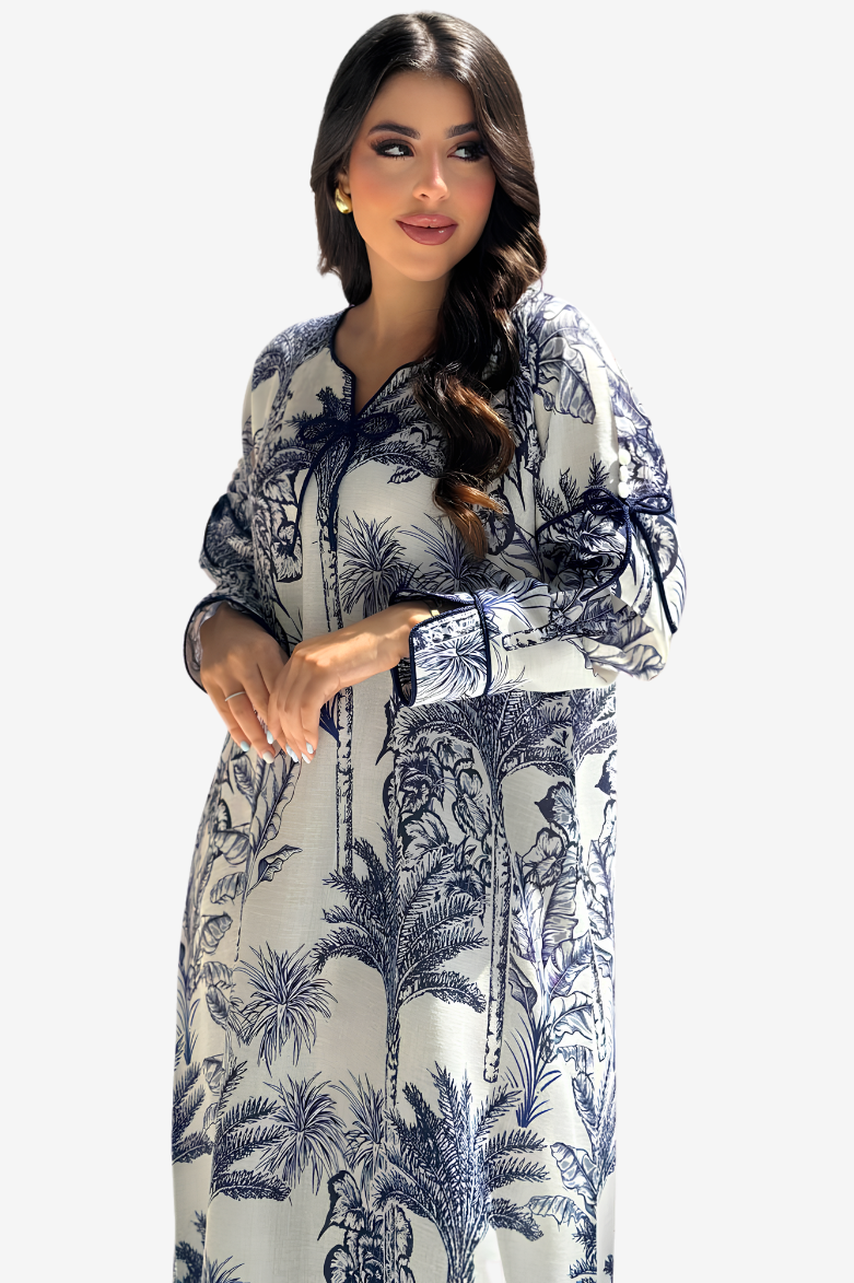 A woman modeling a long-sleeved kaftan dress with a blue and navy botanical print on a white background, set against a plain light gray studio backdrop.