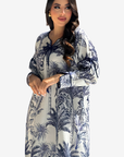 A woman modeling a long-sleeved kaftan dress with a blue and navy botanical print on a white background, set against a plain light gray studio backdrop.