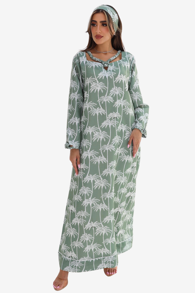 A woman modeling a long-sleeve maxi dress with a white palm tree print on a sage green background, paired with a matching green headband, against a plain white studio background.
