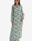 A woman modeling a long-sleeve maxi dress with a white palm tree print on a sage green background, paired with a matching green headband, against a plain white studio background.