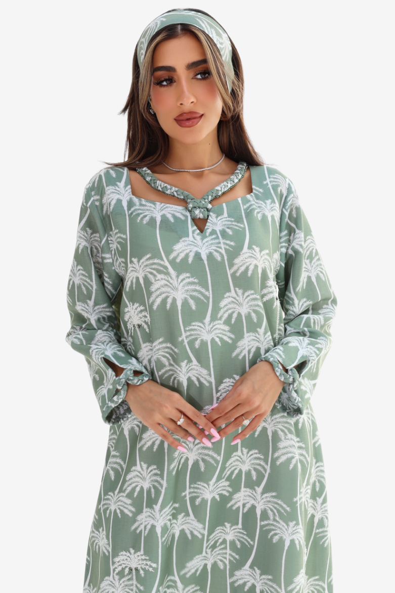 A woman wearing a sage green long-sleeve tunic dress with a white palm tree pattern, styled with a matching headband and jewelry against a plain light gray background.