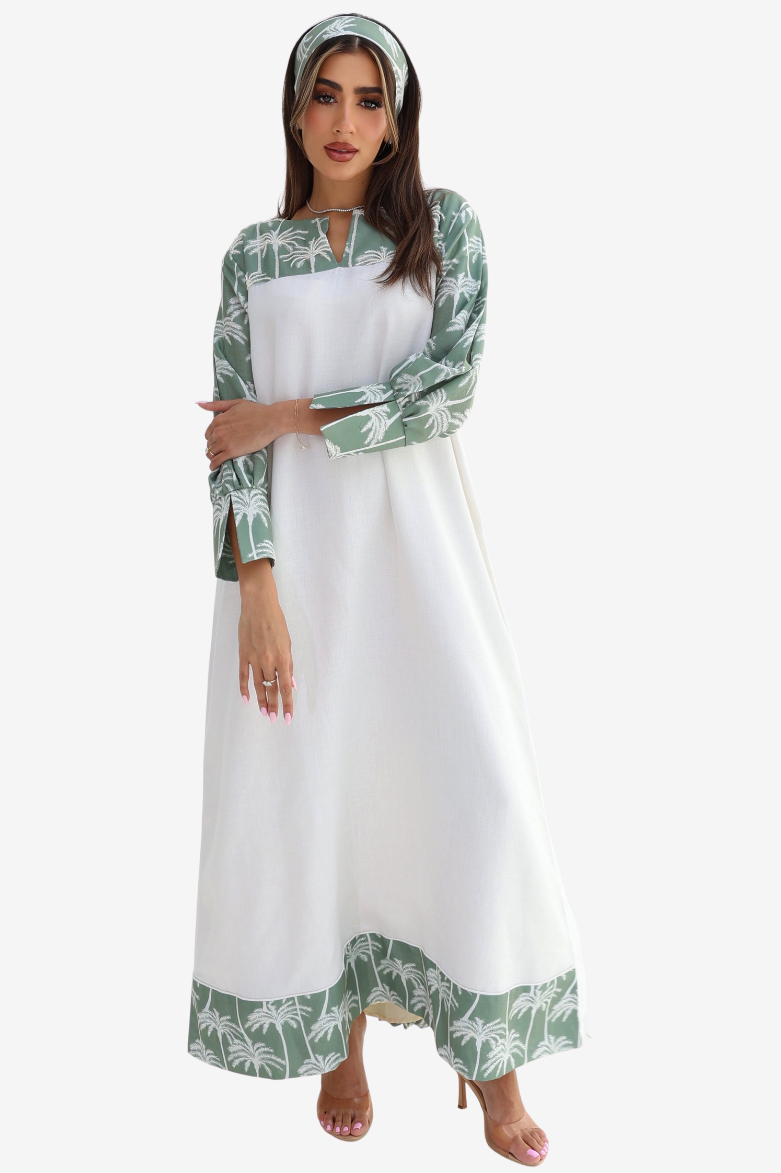 A woman modeling a long white dress with green tropical leaf print on the sleeves, hem, and matching headscarf, set against a plain light gray background.
