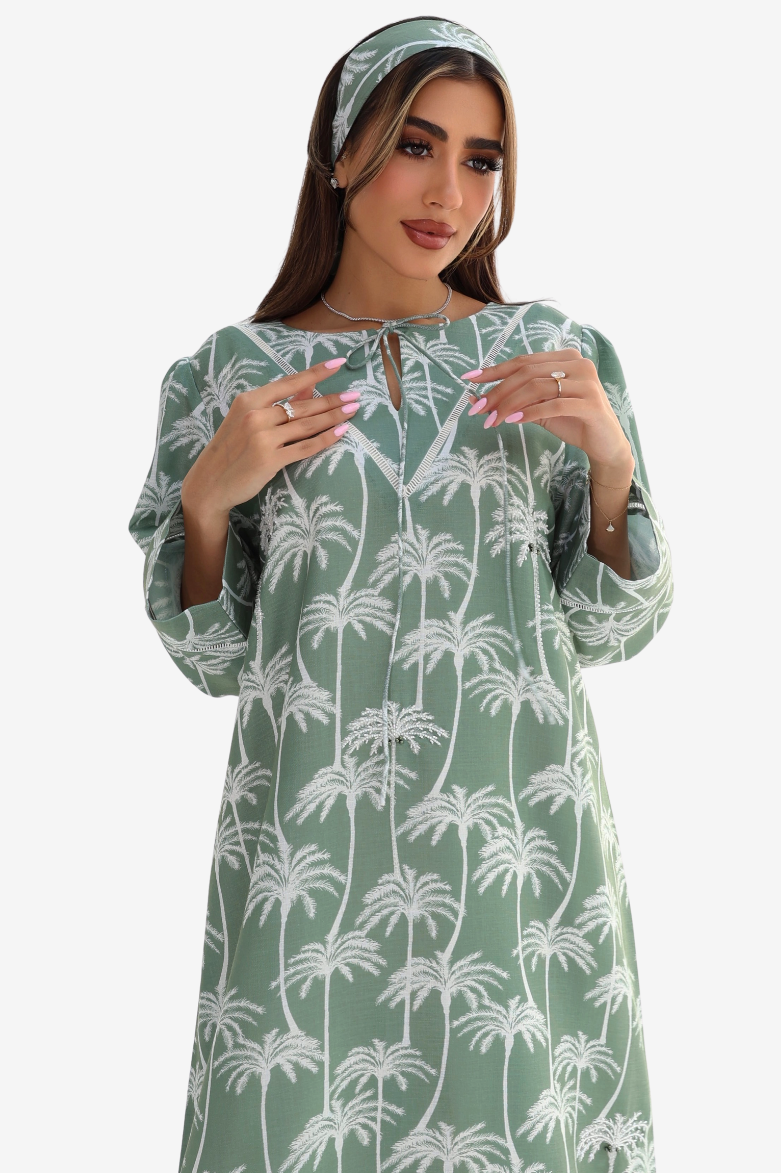 A woman wearing a sage green long-sleeve tunic dress with a white palm tree pattern, paired with a matching headband, set against a plain light gray background.