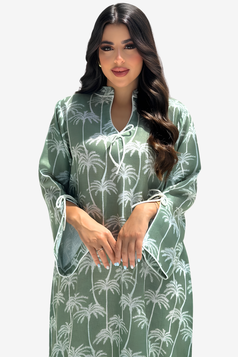 A woman modeling a sage green kaftan dress with a white palm tree pattern, featuring a split neckline with tie details and wide sleeves, set against a plain light gray background.