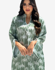 A woman modeling a sage green kaftan dress with a white palm tree pattern, featuring a split neckline with tie details and wide sleeves, set against a plain light gray background.