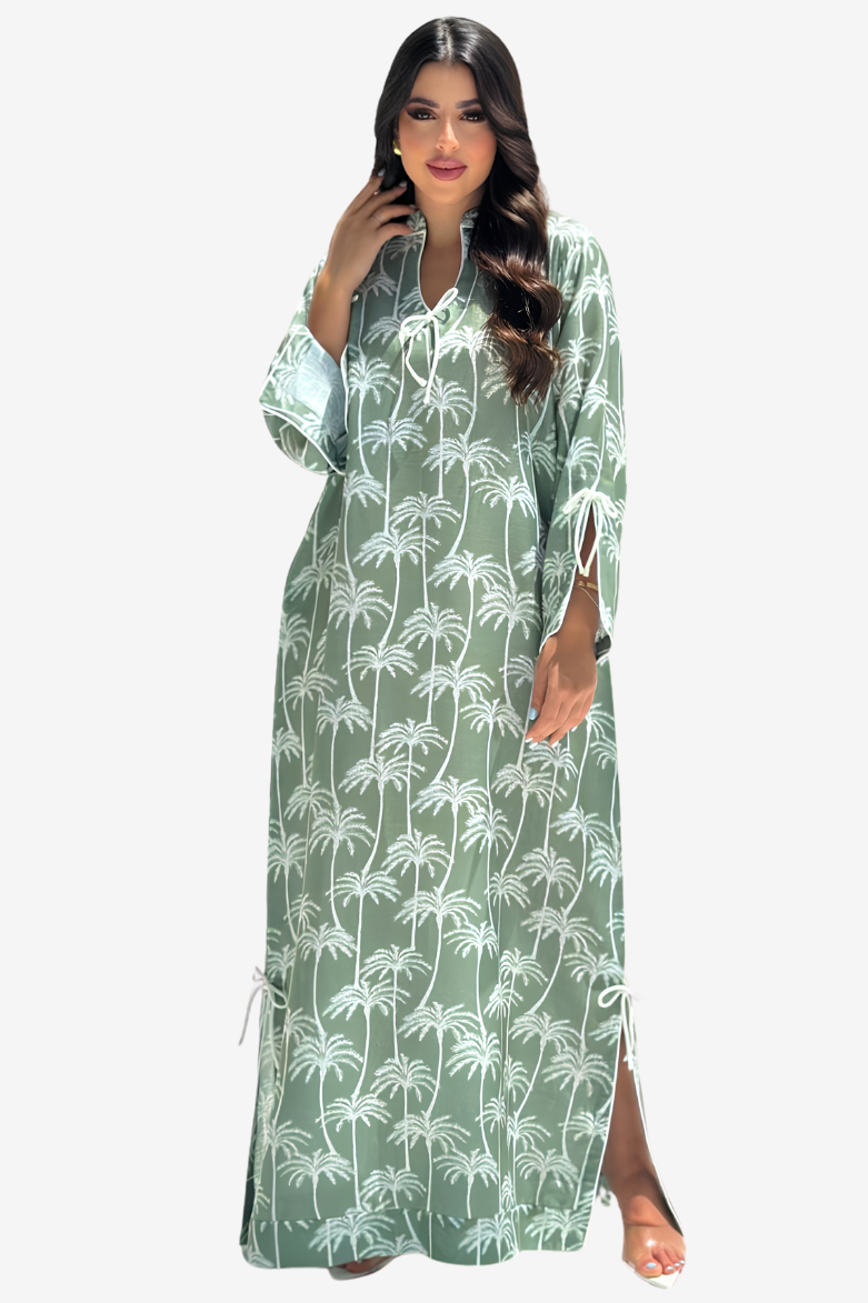 A woman modeling a long-sleeved, floor-length kaftan dress in sage green with a white palm tree print, set against a plain light gray background.