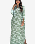 A woman modeling a long-sleeved, floor-length kaftan dress in sage green with a white palm tree print, set against a plain light gray background.