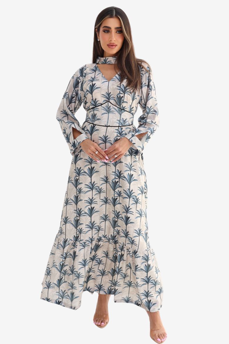 A woman modeling a long-sleeve maxi dress with a blue palm tree print on a cream background, set against a plain light gray studio background.