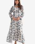 A woman modeling a long-sleeve maxi dress with a blue palm tree print on a cream background, set against a plain light gray studio background.