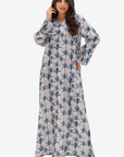 A woman modeling a long-sleeved maxi dress with a blue palm tree pattern on a light beige background, set against a plain light gray studio backdrop.