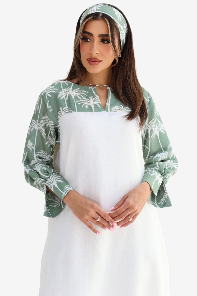 A woman modeling a long-sleeve maternity dress with a sage green and white palm tree print on the sleeves and collar, paired with a solid white front panel and a matching green and white headband.