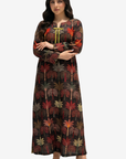 A woman modeling a long-sleeved maxi dress with a dark brown base and an all-over palm tree print in red, green, and beige. The dress features a round neckline with a yellow tie detail at the center chest.