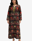 A woman modeling a long-sleeved maxi dress with an all-over palm tree print on a black background, featuring a yellow tie at the neckline, set against a plain light gray studio background.
