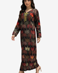 A woman modeling a long-sleeved maxi dress with a tropical palm tree print in red, green, and brown tones on a black background, featuring a yellow tie detail at the neckline, set against a plain light gray background.